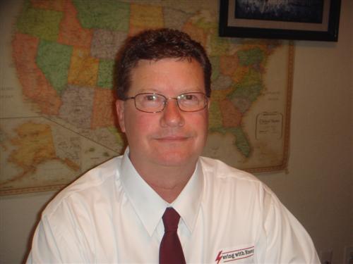 Headshot of a man wearing glasses and a white shirt with a red logo and tie, standing in front of a wall map of the United States.